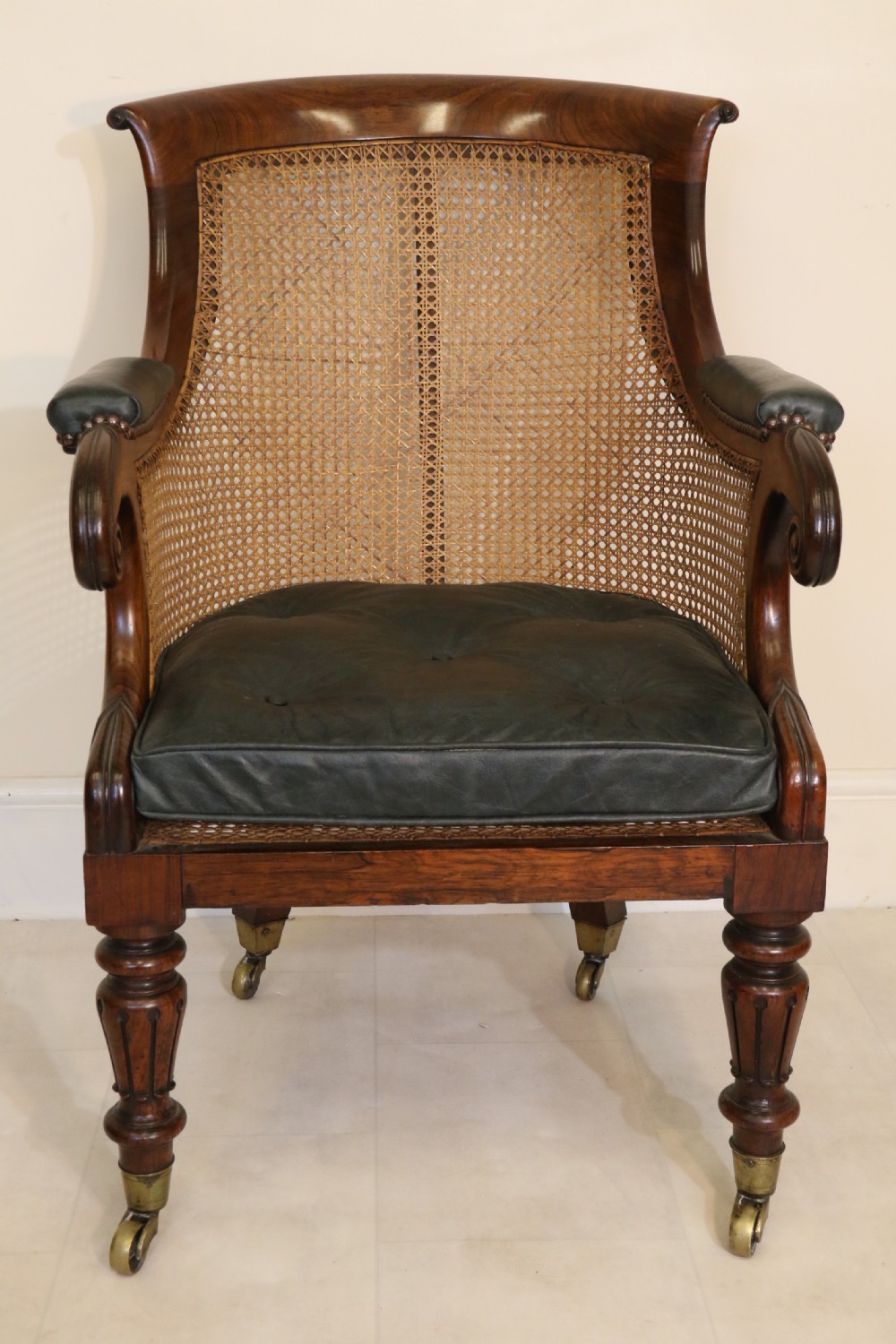 A pair of Regency rosewood bergere library armchairs with hand-woven cane panels, deep green leather cushions, scrolled arms, and turned legs on brass castors. View VI