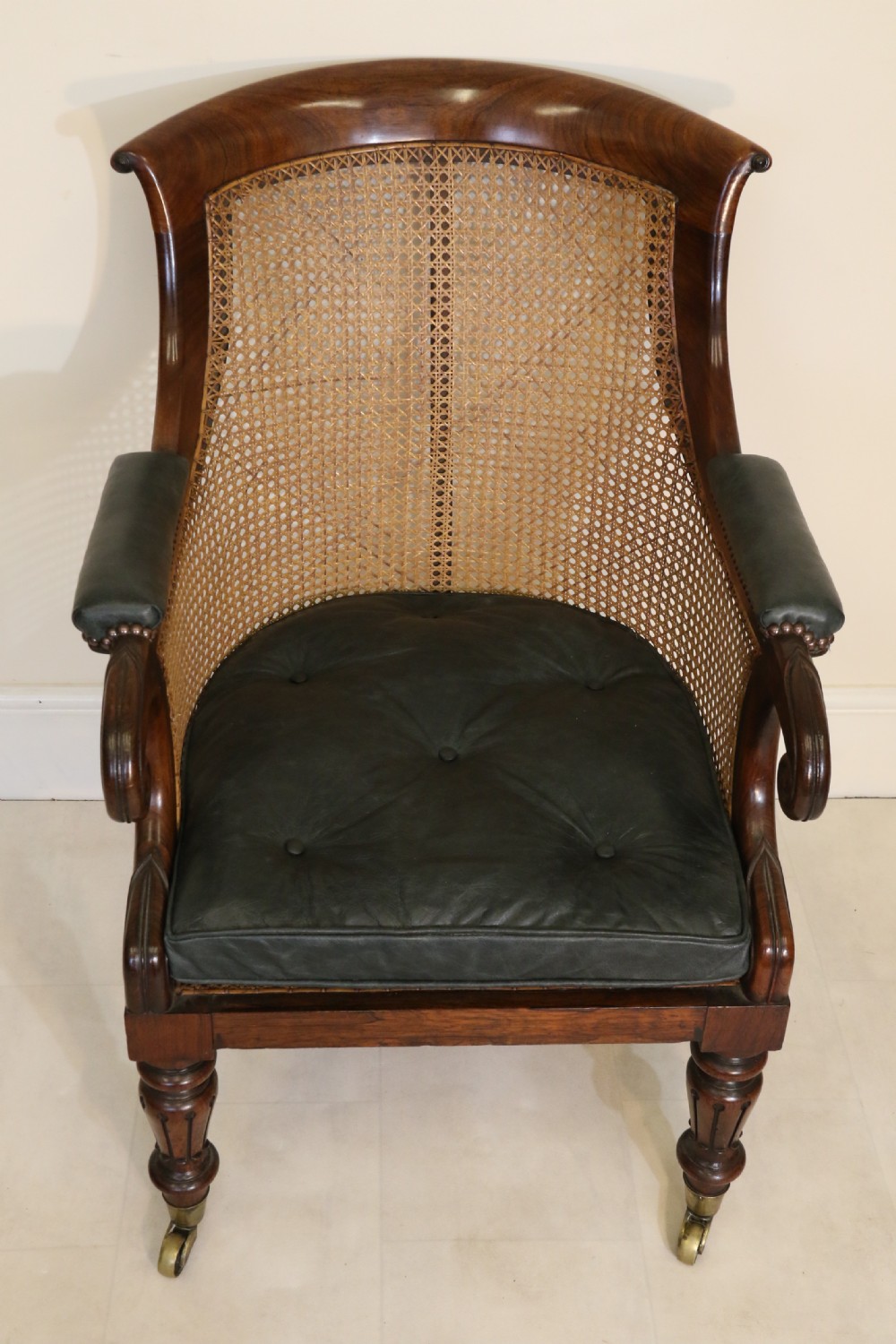 A pair of Regency rosewood bergere library armchairs with hand-woven cane panels, deep green leather cushions, scrolled arms, and turned legs on brass castors. View XVIII