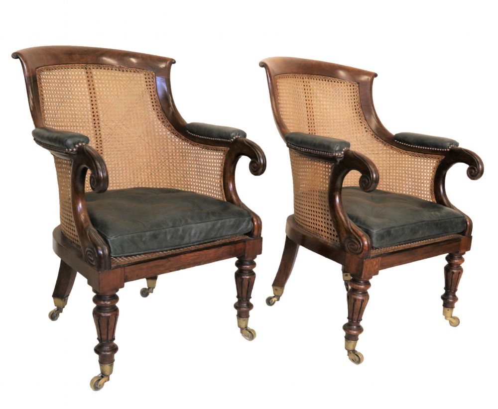 A pair of Regency rosewood bergere library armchairs with hand-woven cane panels, deep green leather cushions, scrolled arms, and turned legs on brass castors.