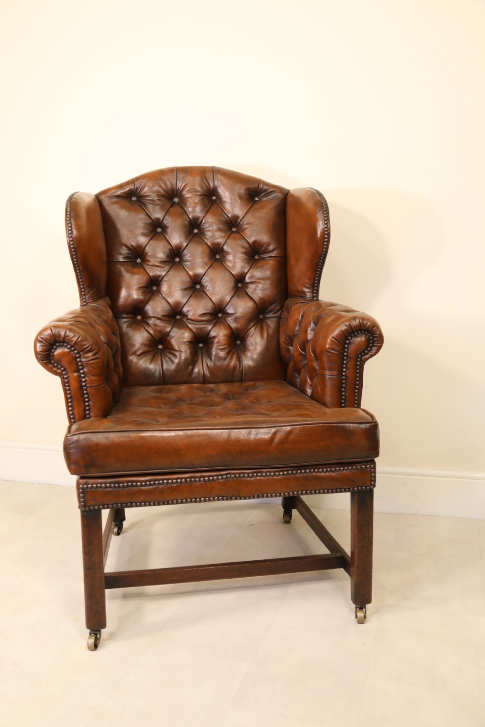 A George III chestnut brown leather wingback armchair featuring deep button tufting, brass stud detail, square wooden legs with stretchers, and brass castors. View II