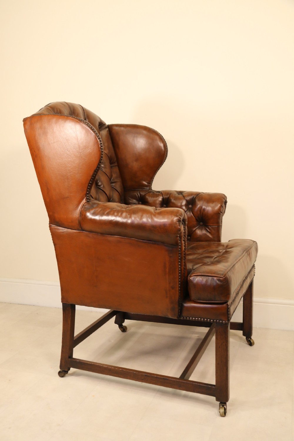 A George III chestnut brown leather wingback armchair featuring deep button tufting, brass stud detail, square wooden legs with stretchers, and brass castors. View III