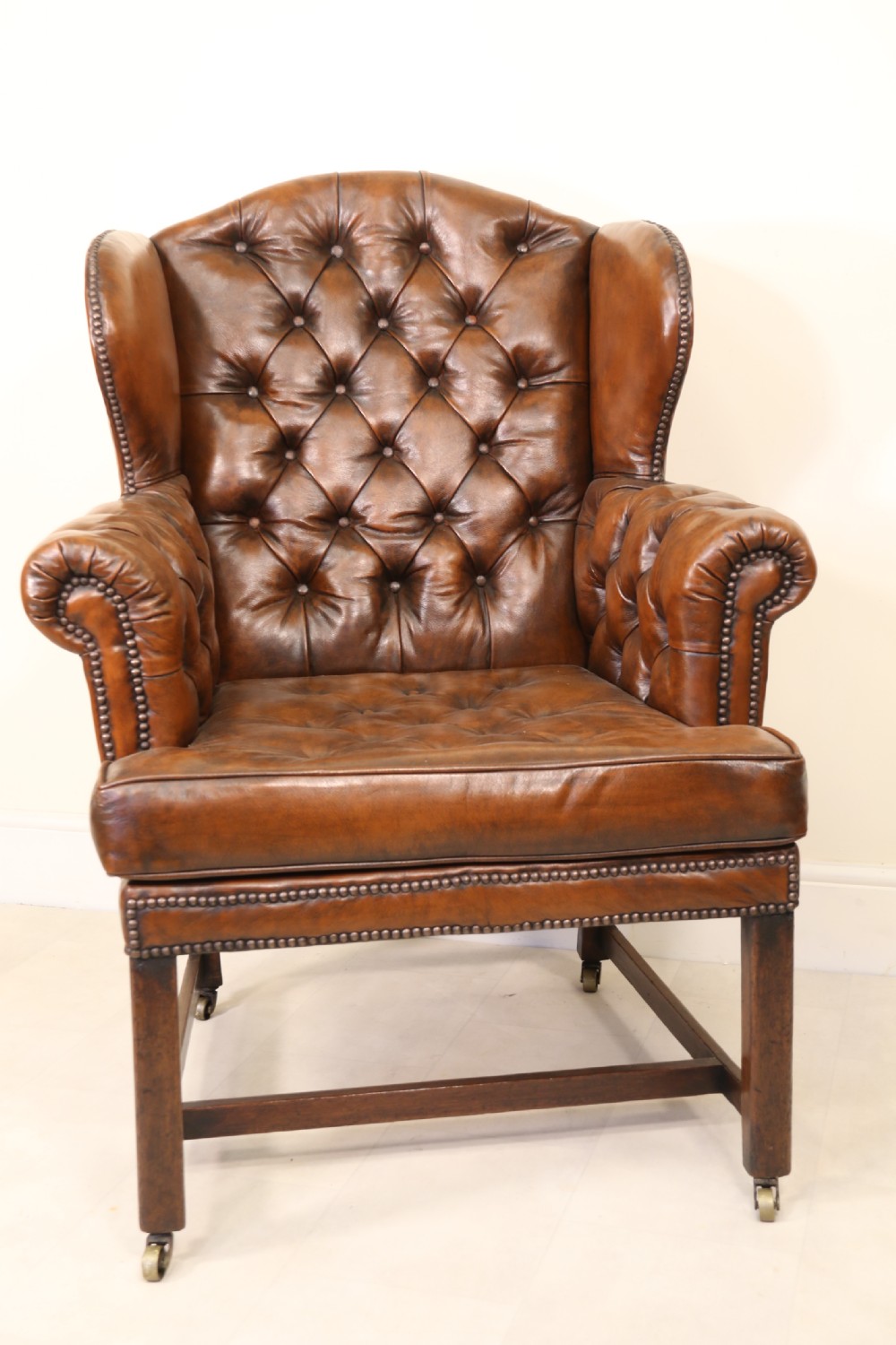 A George III chestnut brown leather wingback armchair featuring deep button tufting, brass stud detail, square wooden legs with stretchers, and brass castors. View IV