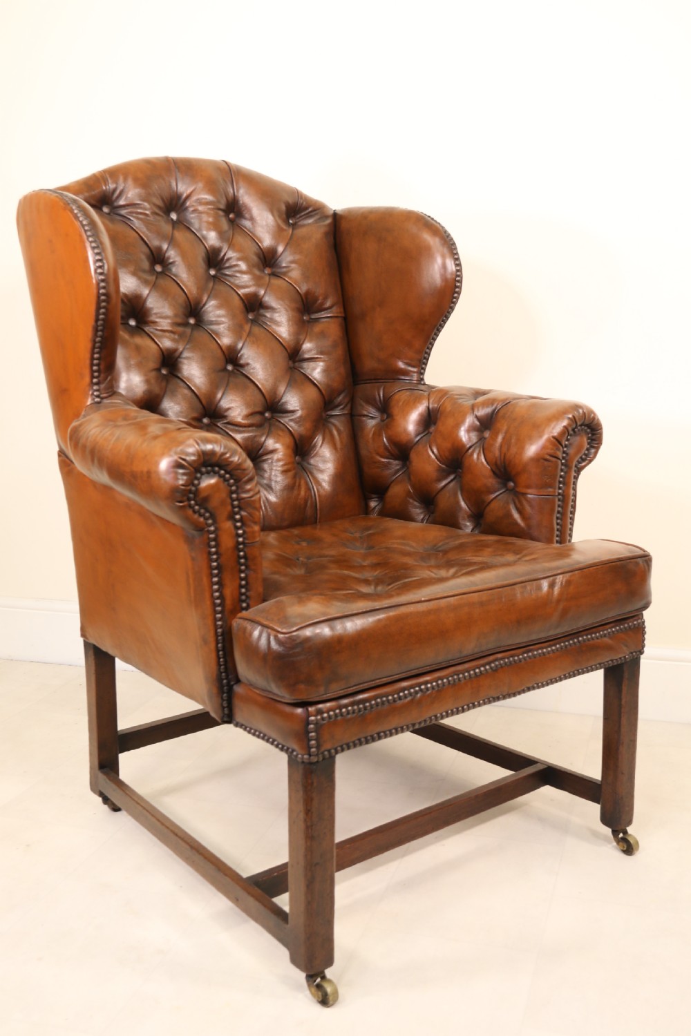 A George III chestnut brown leather wingback armchair featuring deep button tufting, brass stud detail, square wooden legs with stretchers, and brass castors. View V