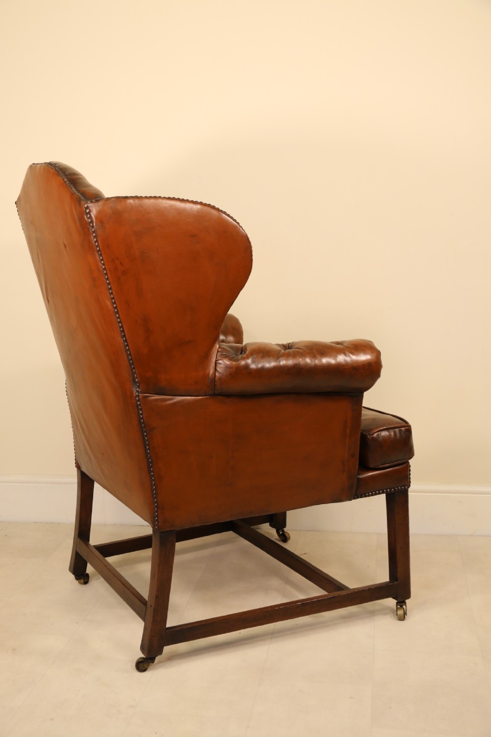 A George III chestnut brown leather wingback armchair featuring deep button tufting, brass stud detail, square wooden legs with stretchers, and brass castors. View VI