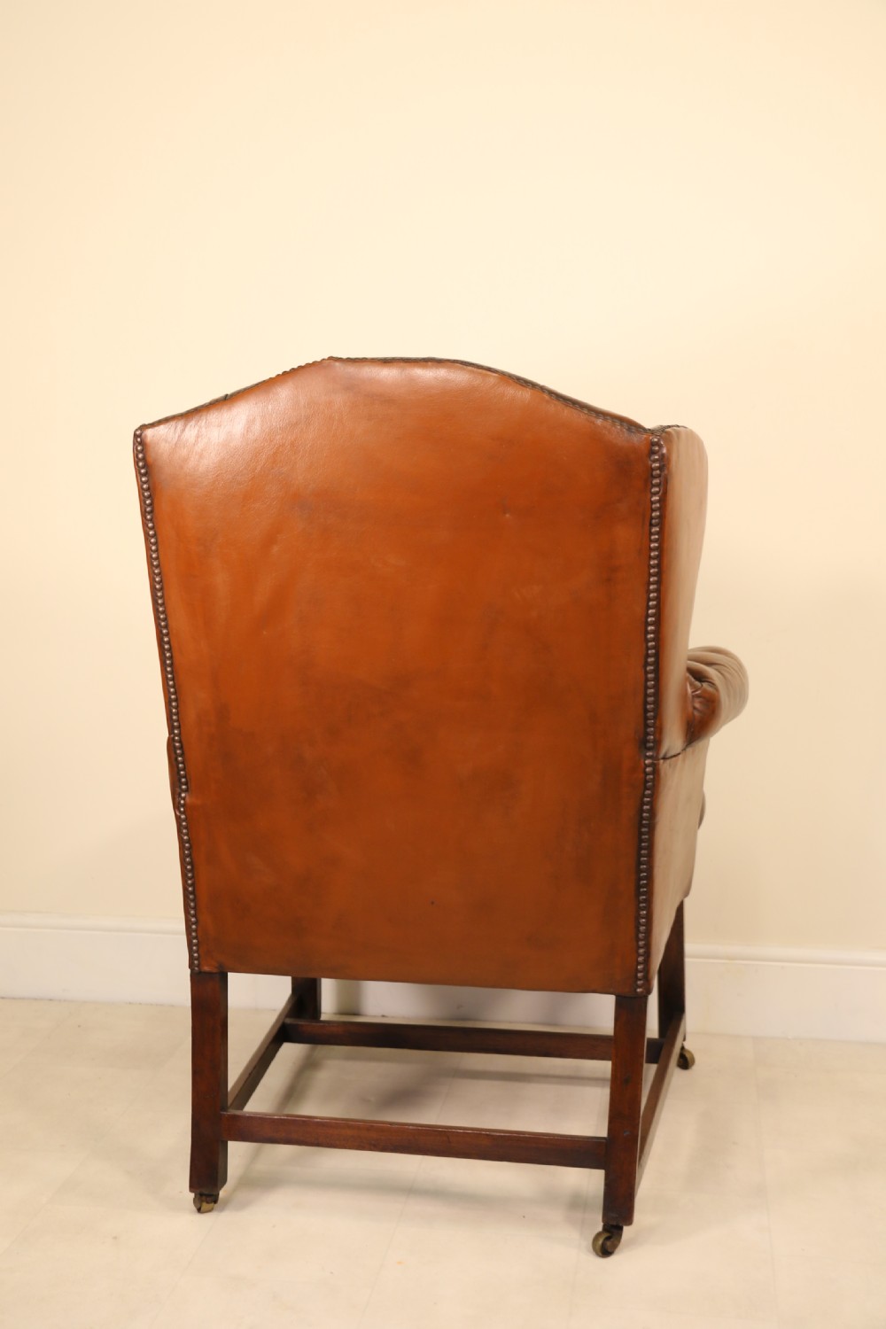 A George III chestnut brown leather wingback armchair featuring deep button tufting, brass stud detail, square wooden legs with stretchers, and brass castors. View VII