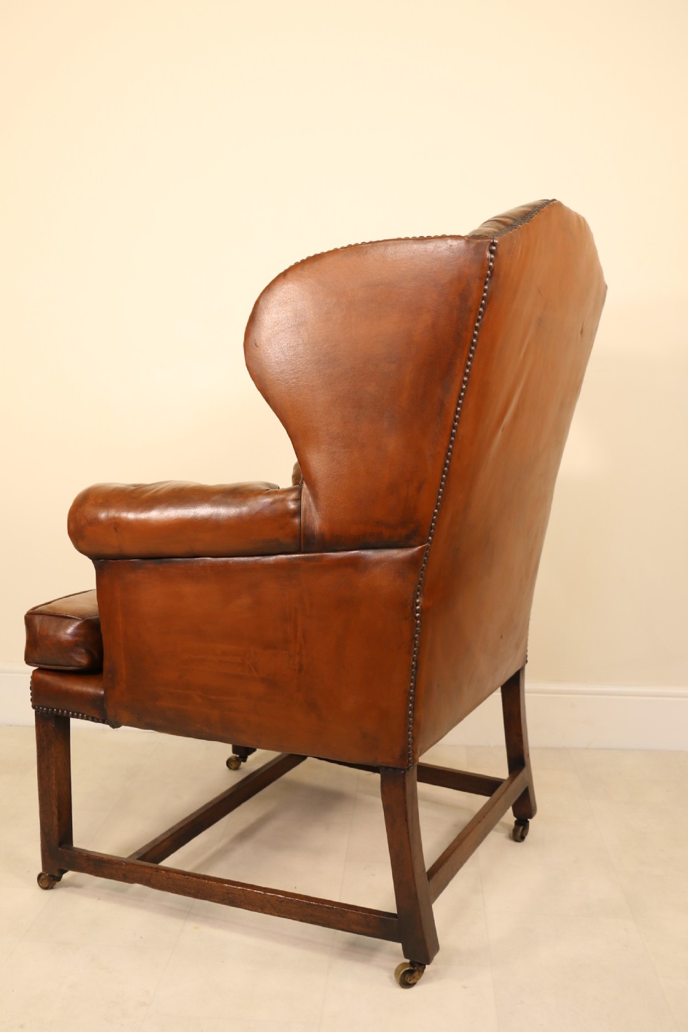 A George III chestnut brown leather wingback armchair featuring deep button tufting, brass stud detail, square wooden legs with stretchers, and brass castors. View VIII