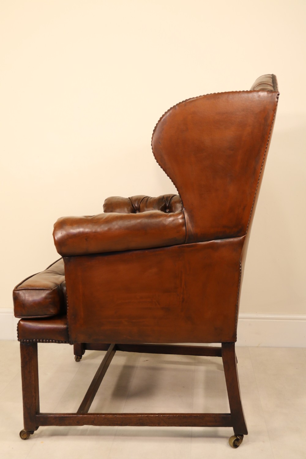 A George III chestnut brown leather wingback armchair featuring deep button tufting, brass stud detail, square wooden legs with stretchers, and brass castors. View IX