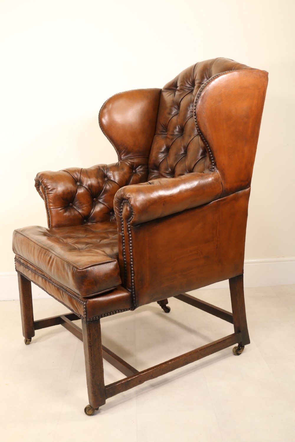 A George III chestnut brown leather wingback armchair featuring deep button tufting, brass stud detail, square wooden legs with stretchers, and brass castors. View X