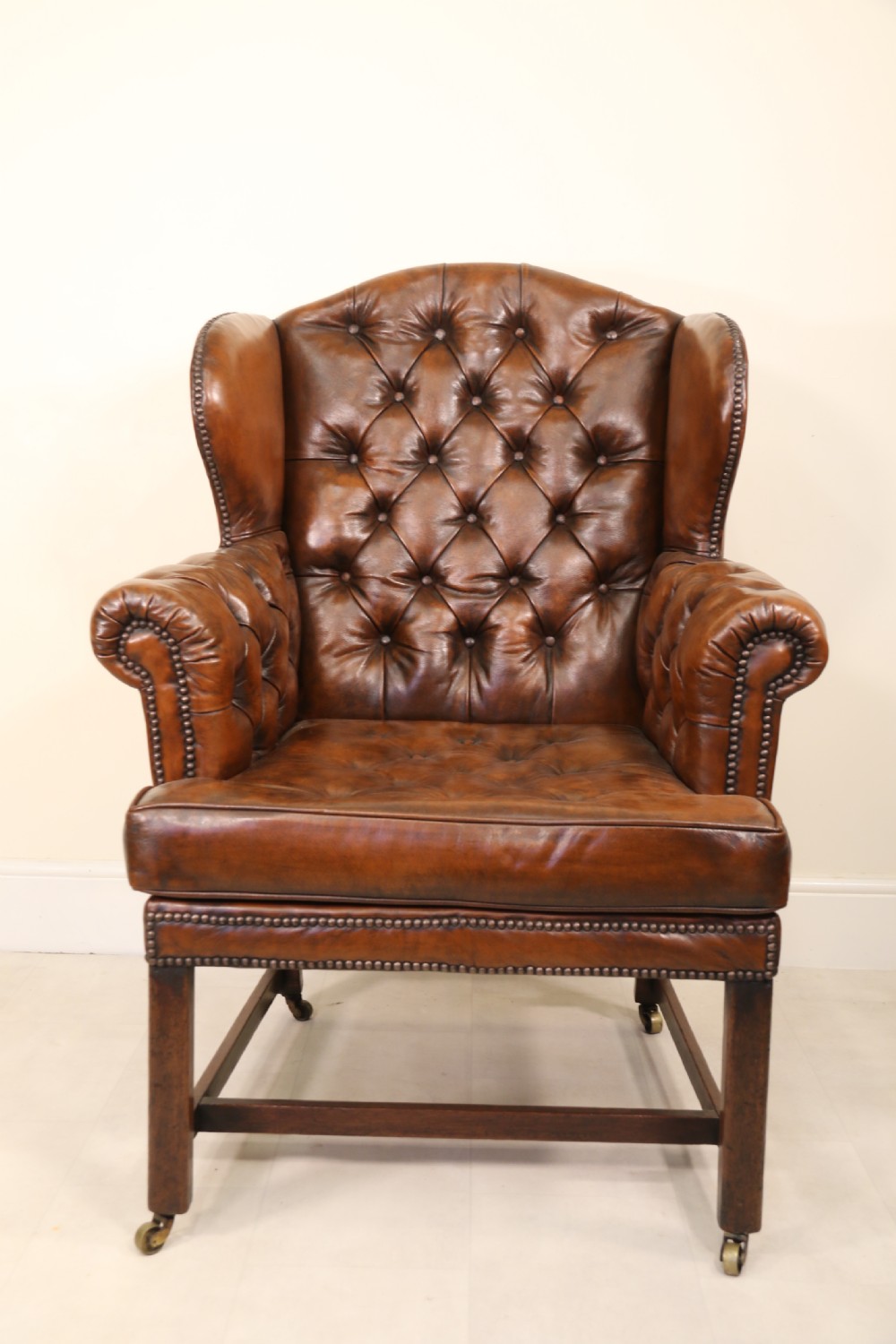 A George III chestnut brown leather wingback armchair featuring deep button tufting, brass stud detail, square wooden legs with stretchers, and brass castors. View XI