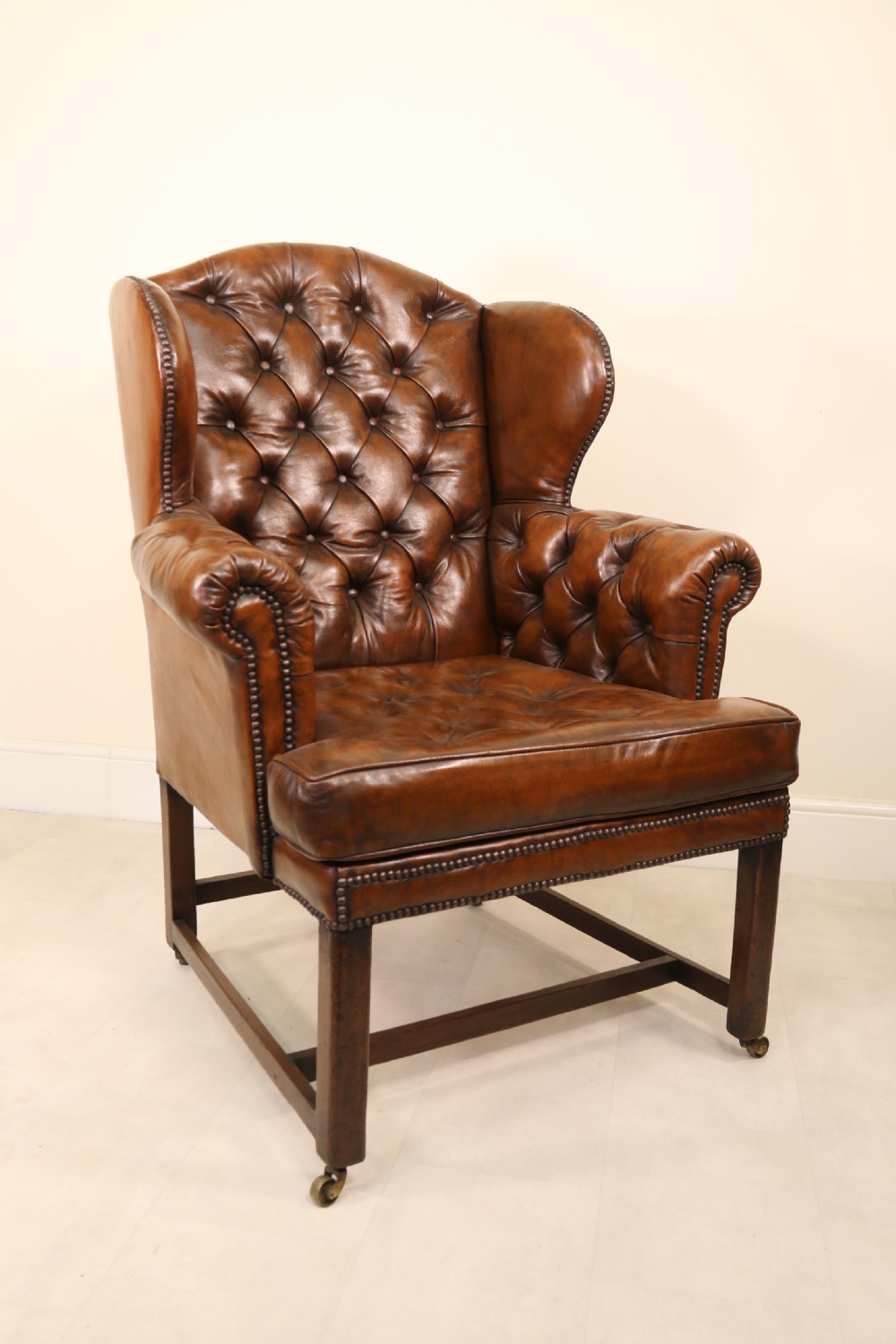 A George III chestnut brown leather wingback armchair featuring deep button tufting, brass stud detail, square wooden legs with stretchers, and brass castors. View XII