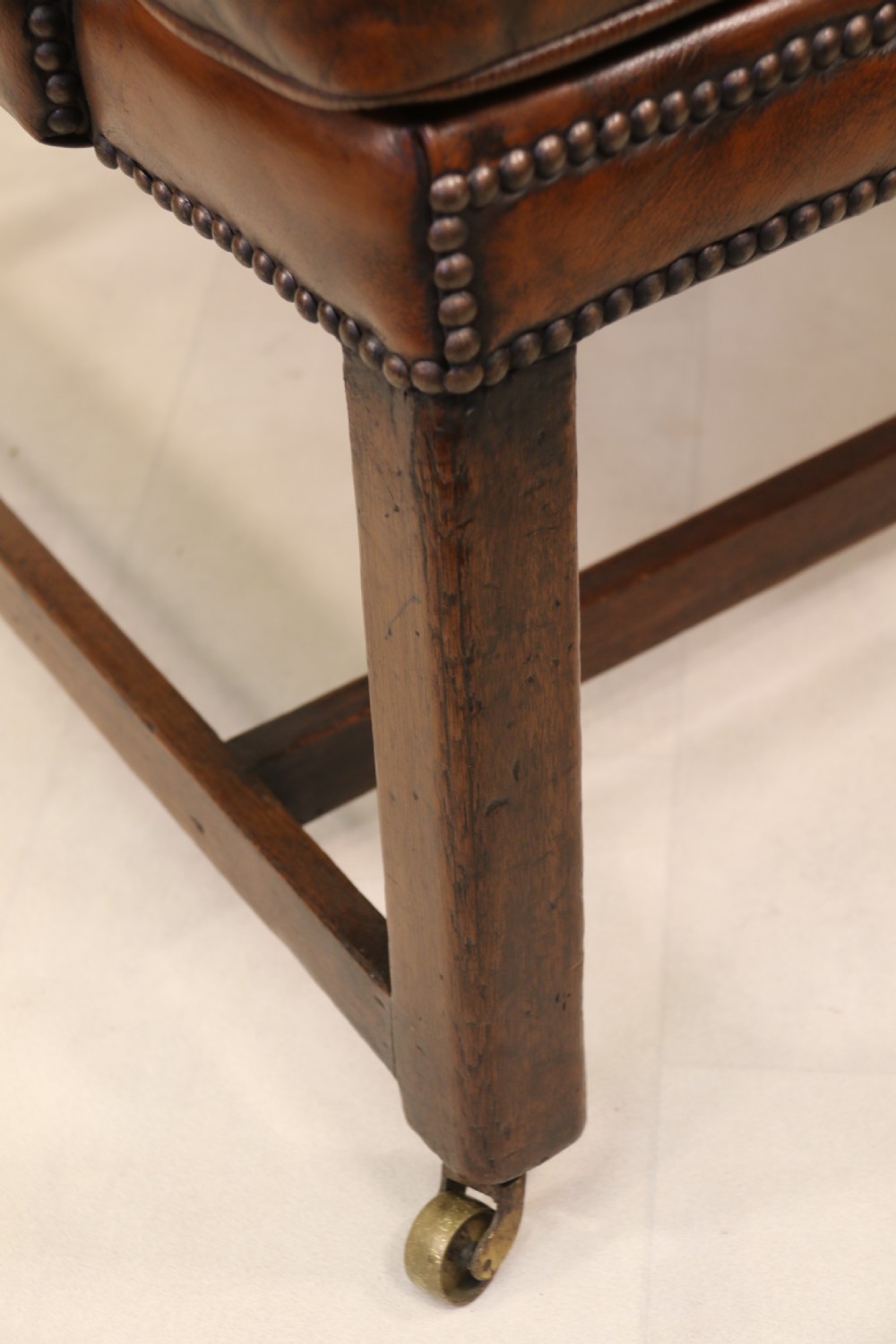 A George III chestnut brown leather wingback armchair featuring deep button tufting, brass stud detail, square wooden legs with stretchers, and brass castors. View XIV