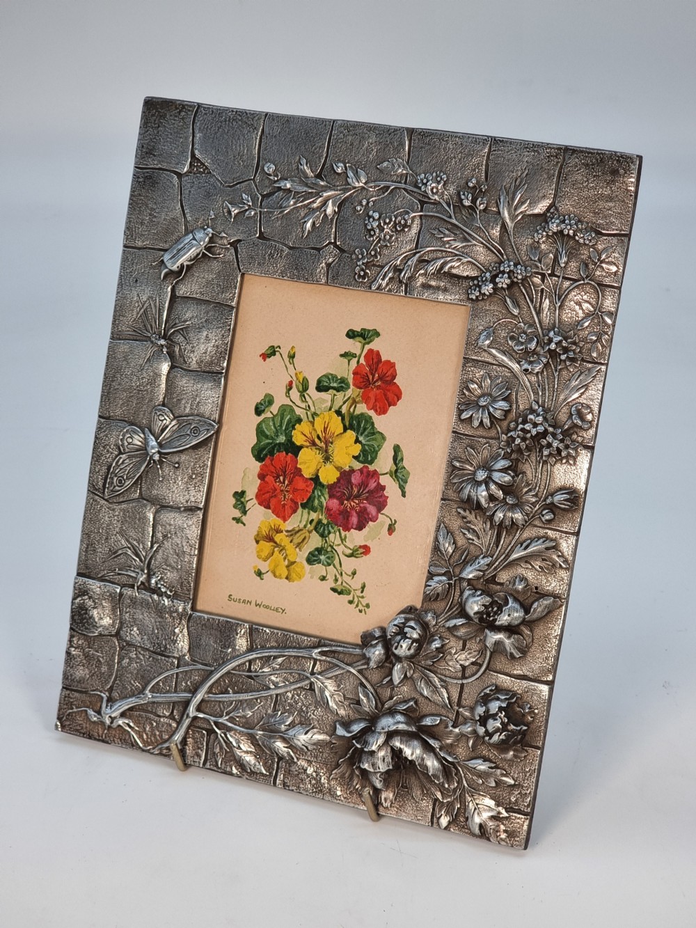 An Aesthetic Movement pewter picture frame featuring embossed insect and floral relief decoration around a central floral watercolour illustration.