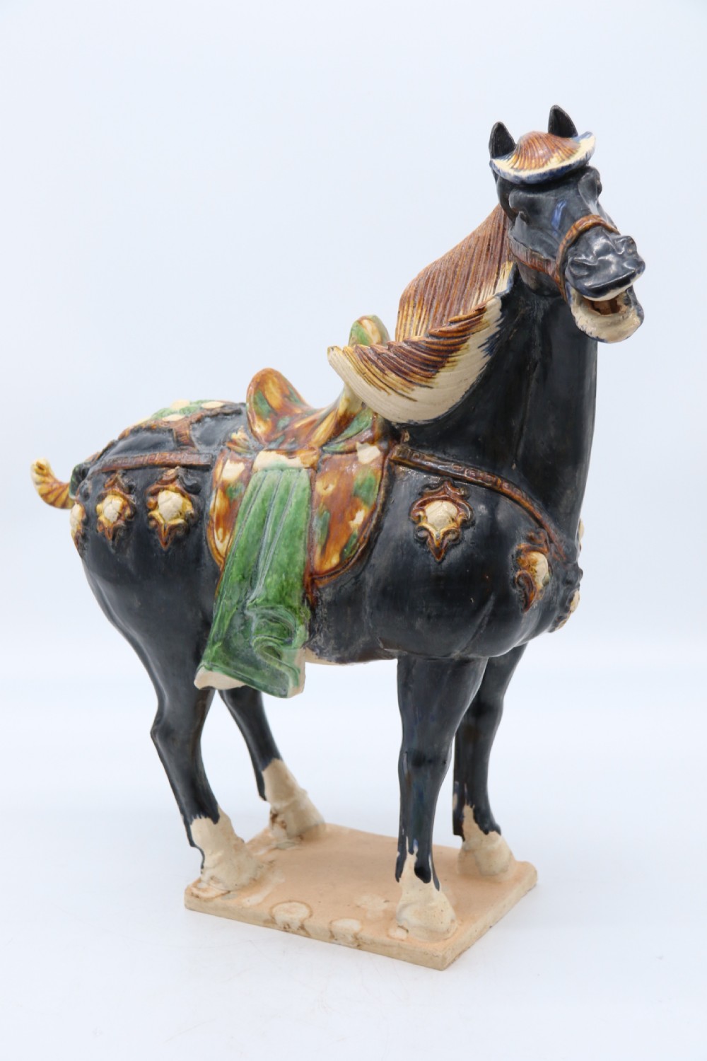 A ceramic Tang Dynasty style horse statue featuring a dark body glaze, amber mane, green draped saddle, and cream base.