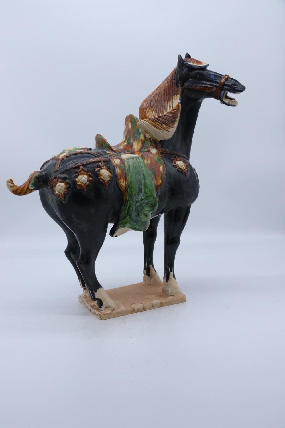 A ceramic Tang Dynasty style horse statue featuring a dark body glaze, amber mane, green draped saddle, and cream base. View II