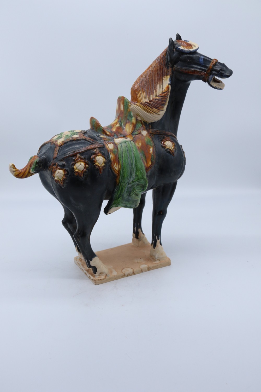 A ceramic Tang Dynasty style horse statue featuring a dark body glaze, amber mane, green draped saddle, and cream base. View III