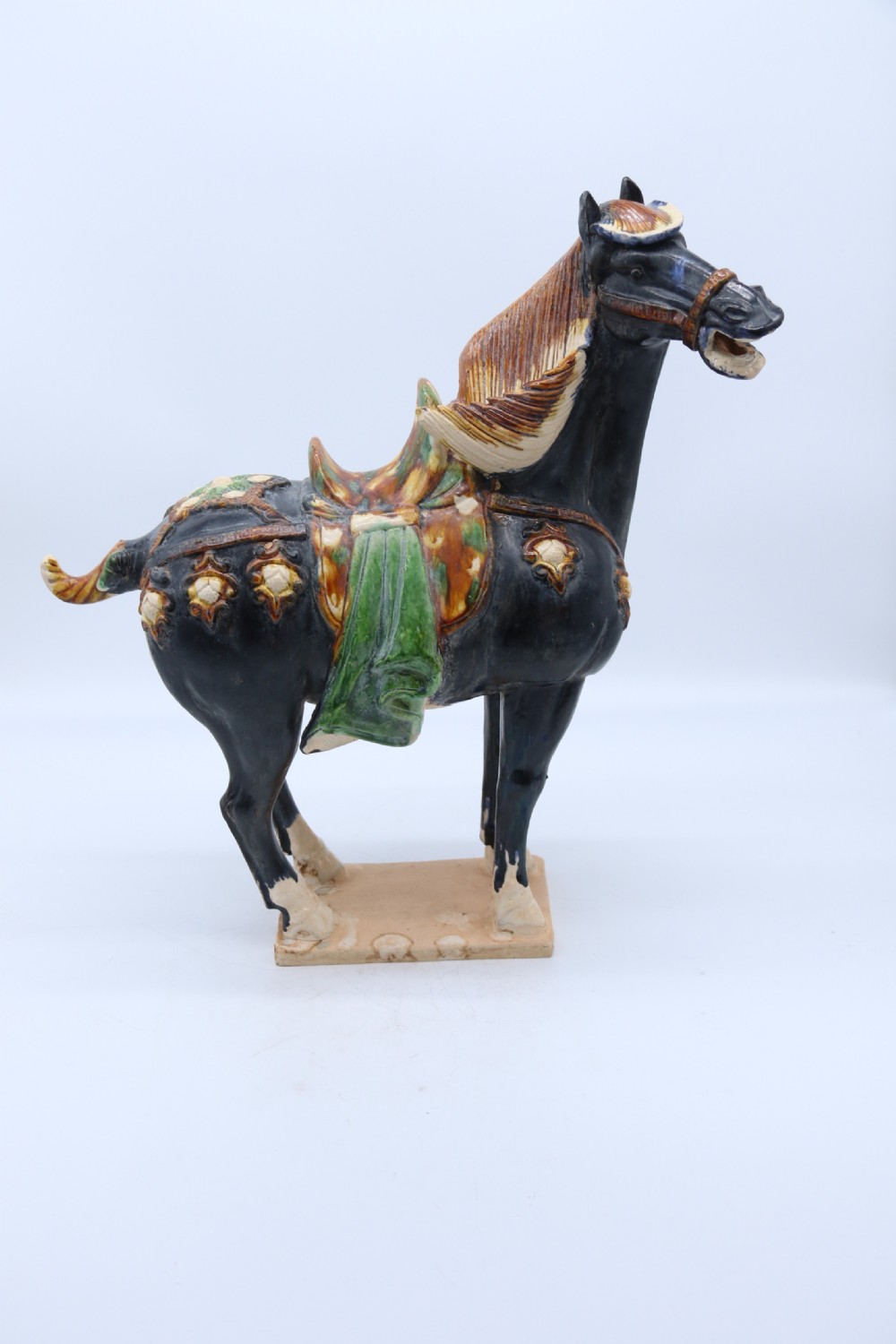 A ceramic Tang Dynasty style horse statue featuring a dark body glaze, amber mane, green draped saddle, and cream base. View IV
