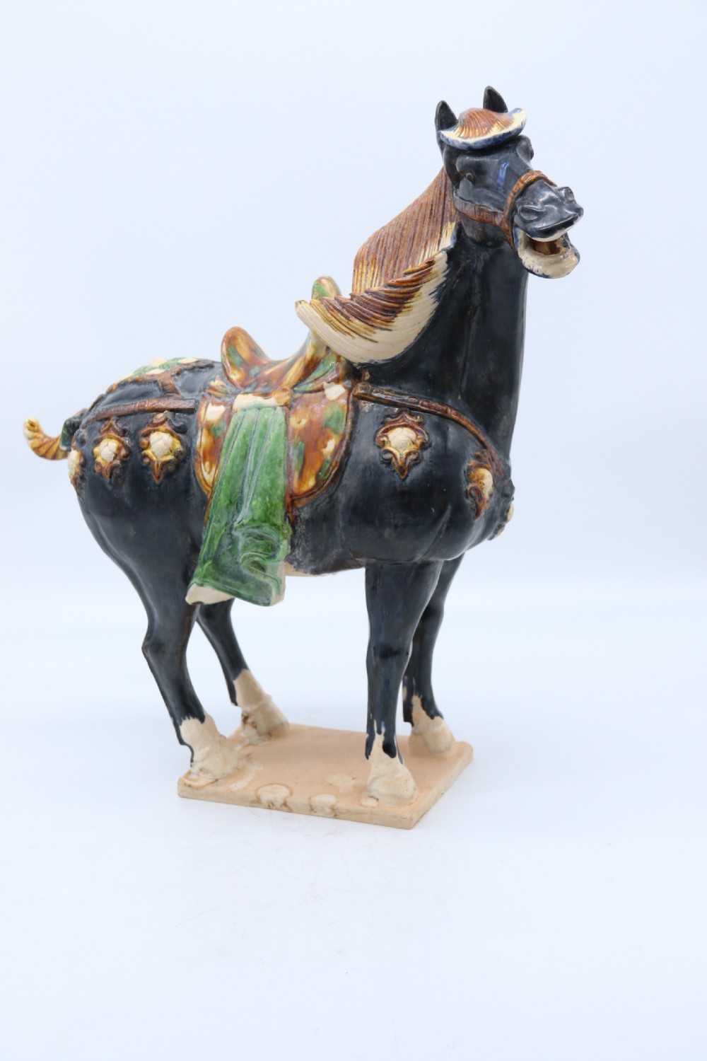 A ceramic Tang Dynasty style horse statue featuring a dark body glaze, amber mane, green draped saddle, and cream base. View V