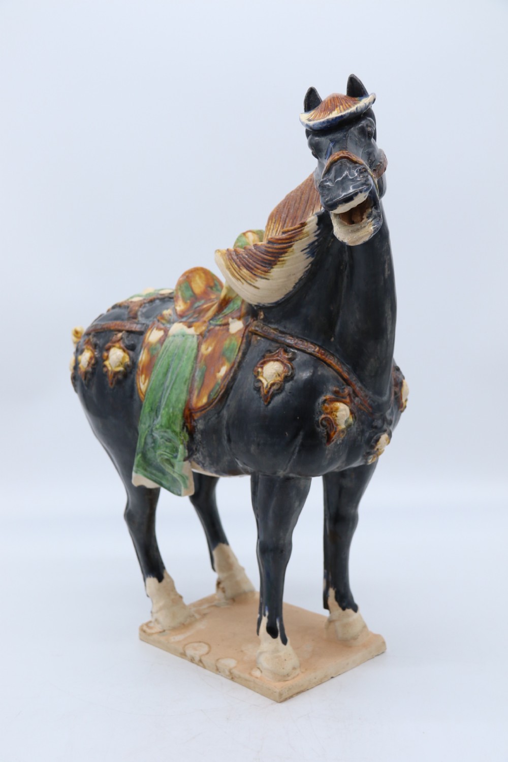 A ceramic Tang Dynasty style horse statue featuring a dark body glaze, amber mane, green draped saddle, and cream base. View VI
