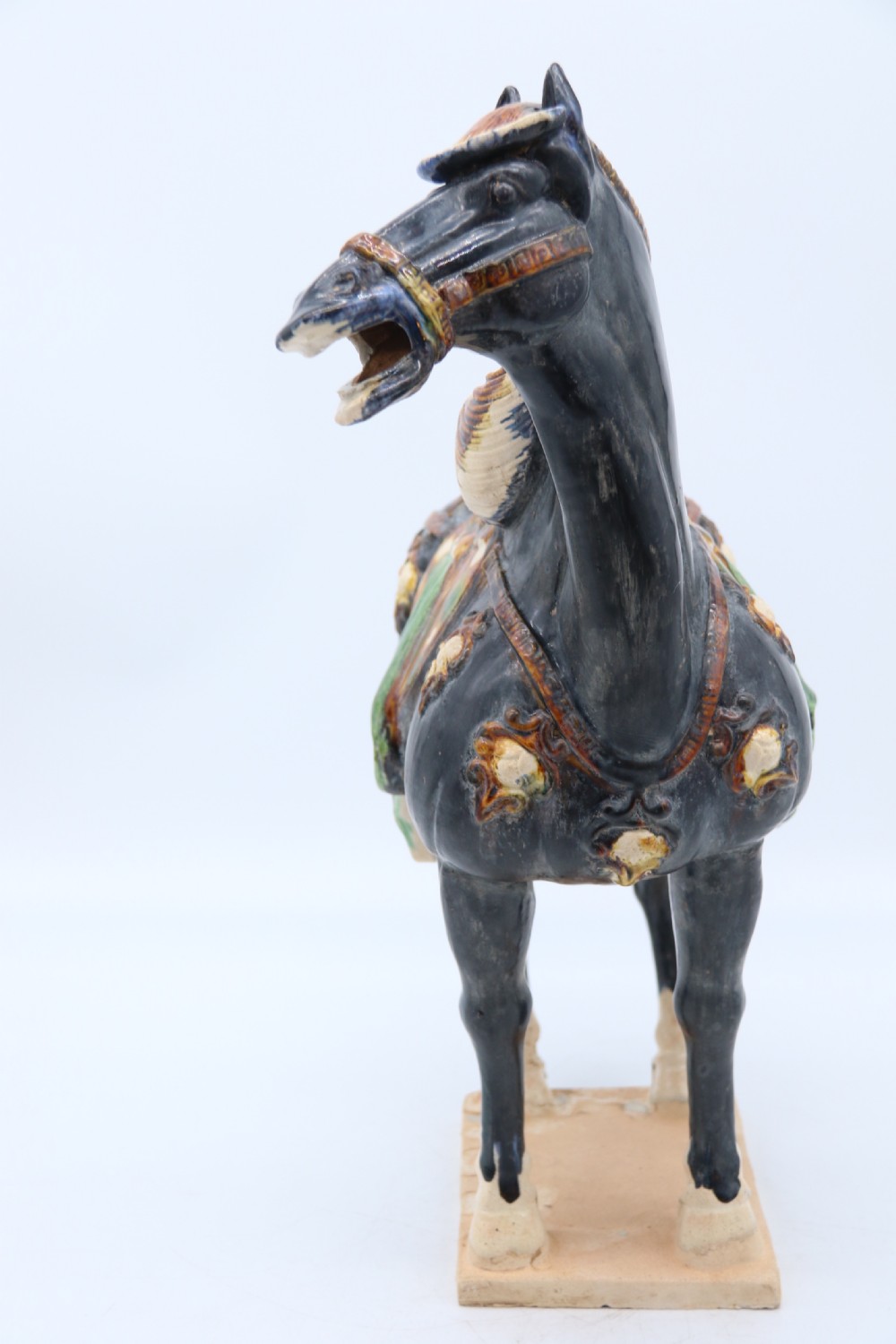 A ceramic Tang Dynasty style horse statue featuring a dark body glaze, amber mane, green draped saddle, and cream base. View VII
