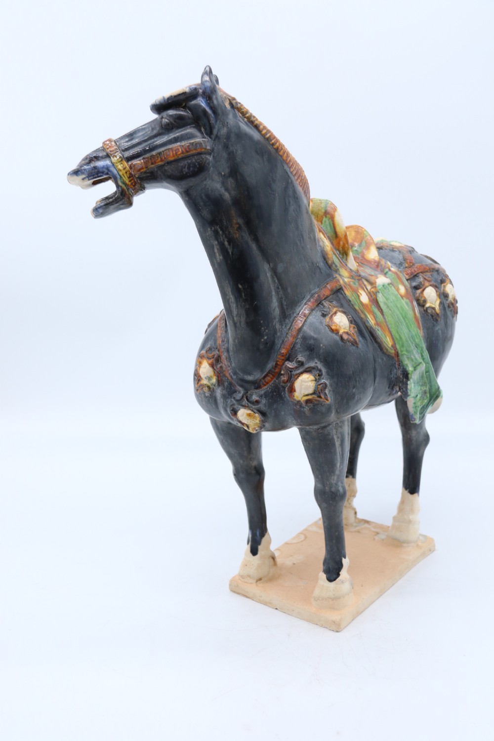 A ceramic Tang Dynasty style horse statue featuring a dark body glaze, amber mane, green draped saddle, and cream base. View VIII
