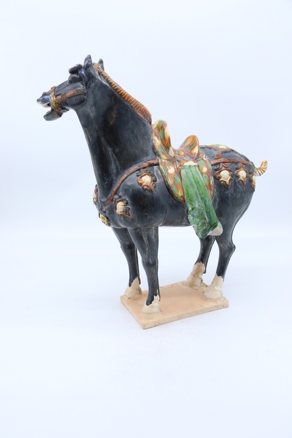 A ceramic Tang Dynasty style horse statue featuring a dark body glaze, amber mane, green draped saddle, and cream base. View IX