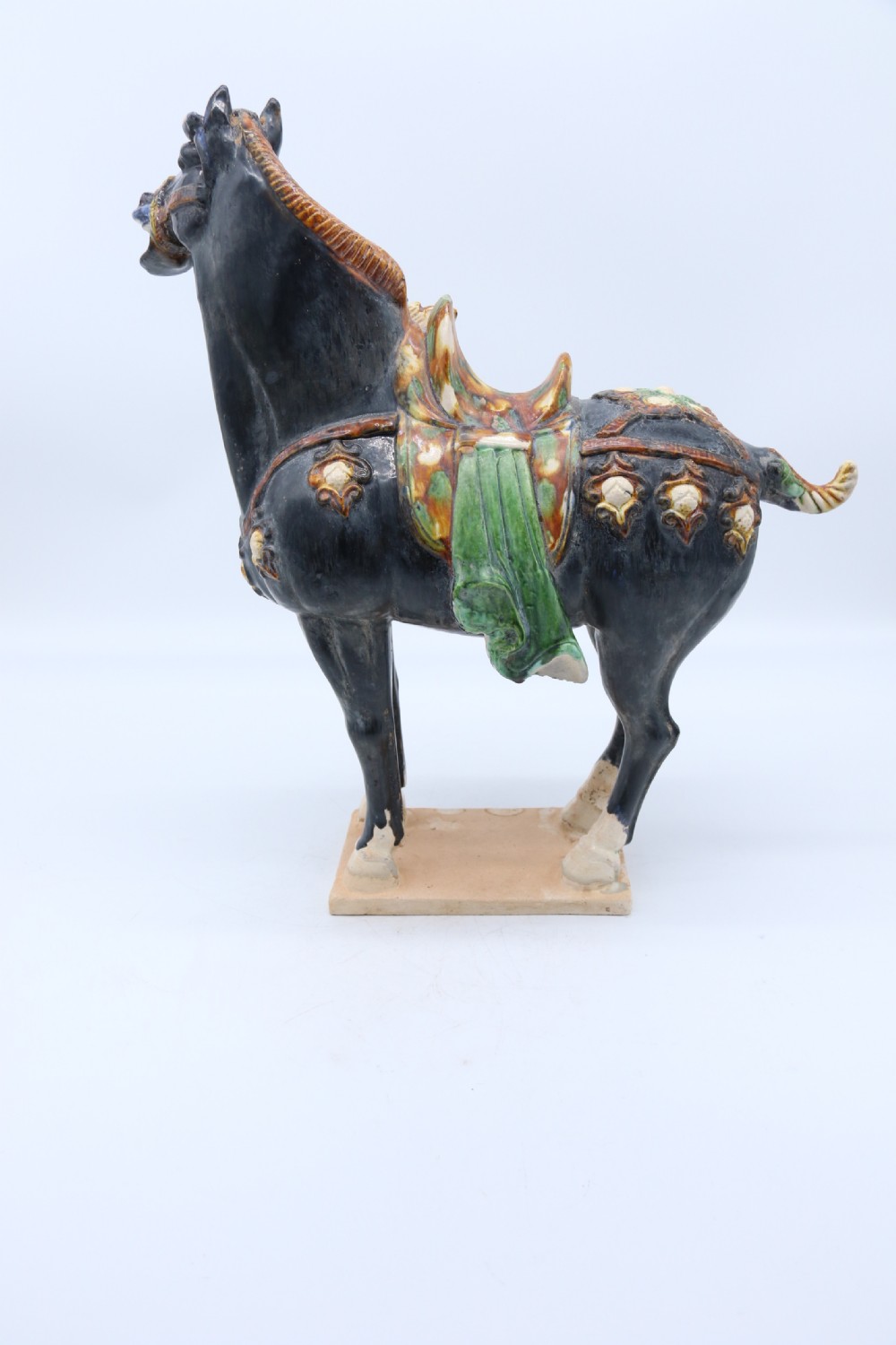 A ceramic Tang Dynasty style horse statue featuring a dark body glaze, amber mane, green draped saddle, and cream base. View X