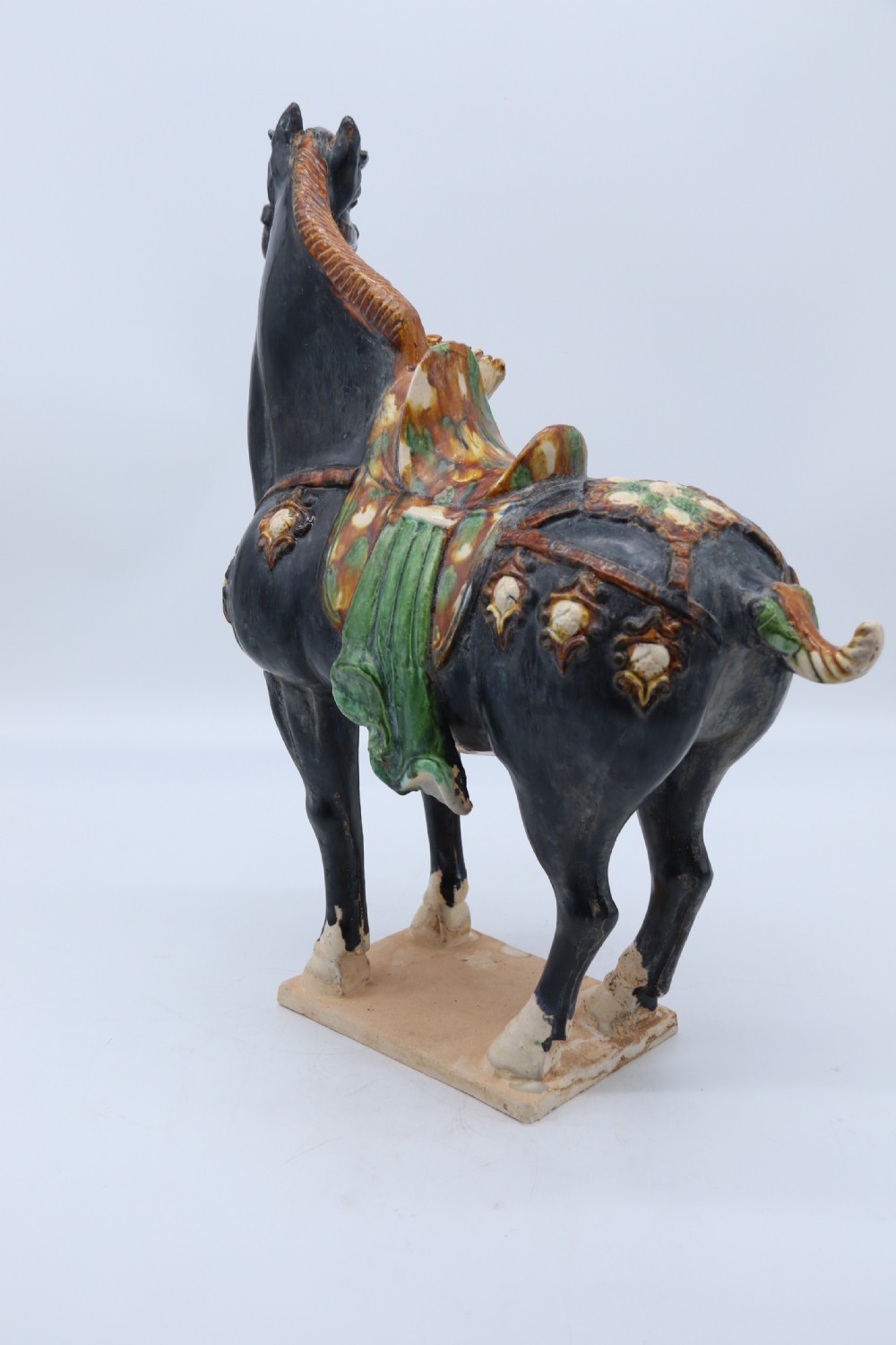 A ceramic Tang Dynasty style horse statue featuring a dark body glaze, amber mane, green draped saddle, and cream base. View XI