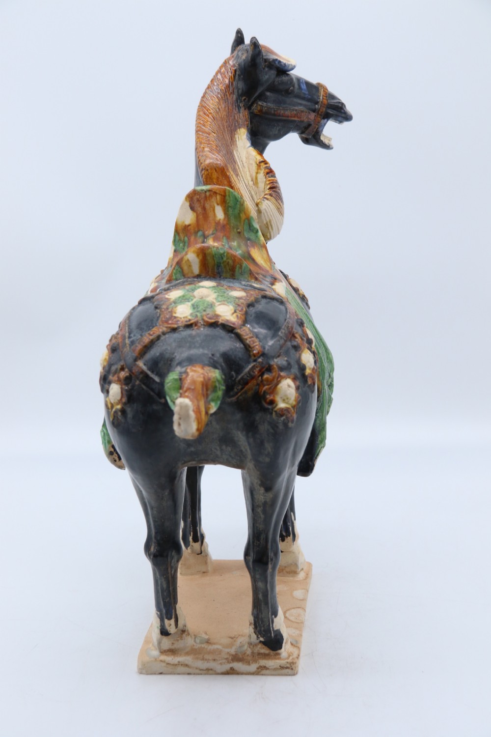 A ceramic Tang Dynasty style horse statue featuring a dark body glaze, amber mane, green draped saddle, and cream base. View XII