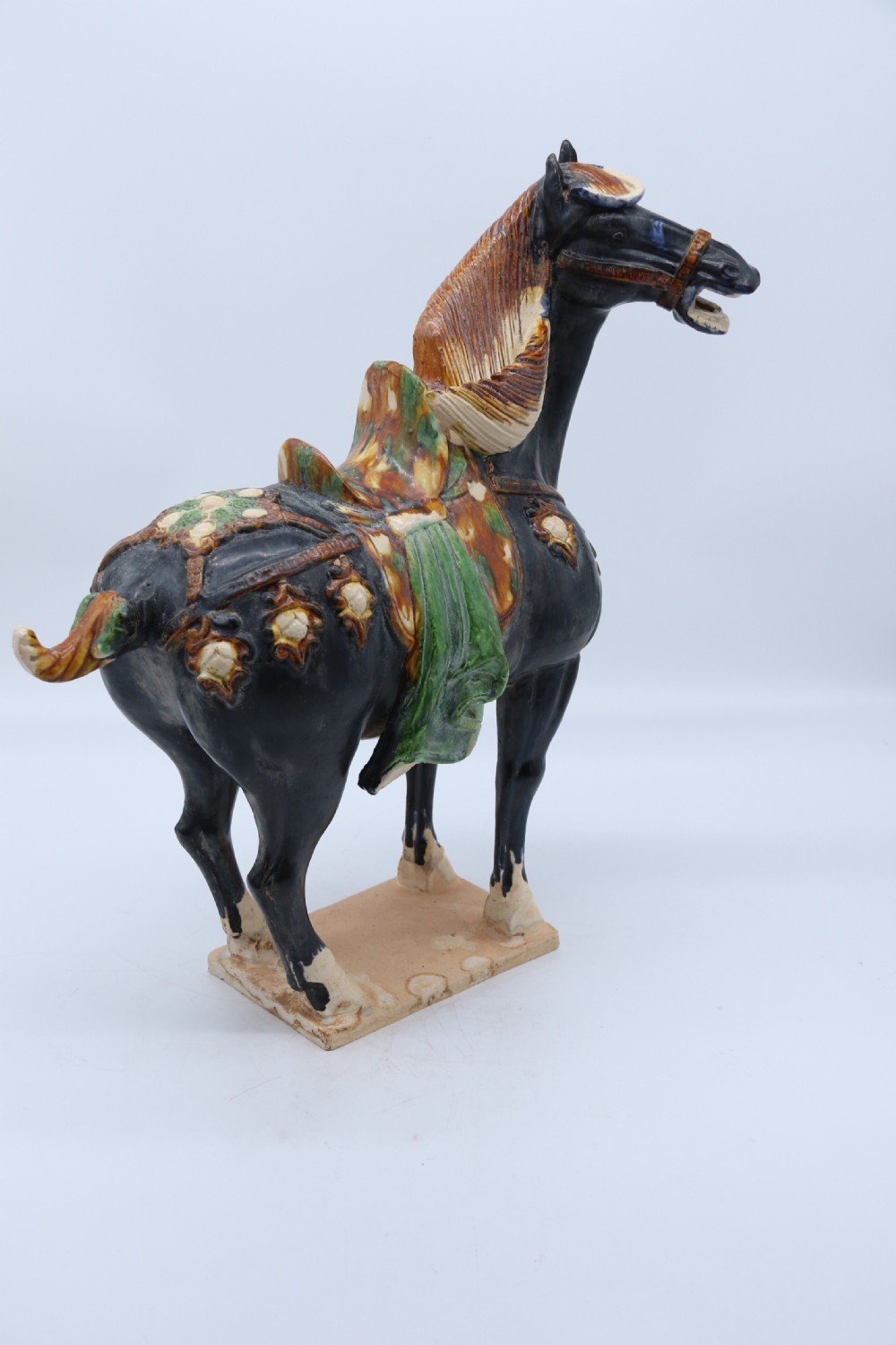 A ceramic Tang Dynasty style horse statue featuring a dark body glaze, amber mane, green draped saddle, and cream base. View XIII