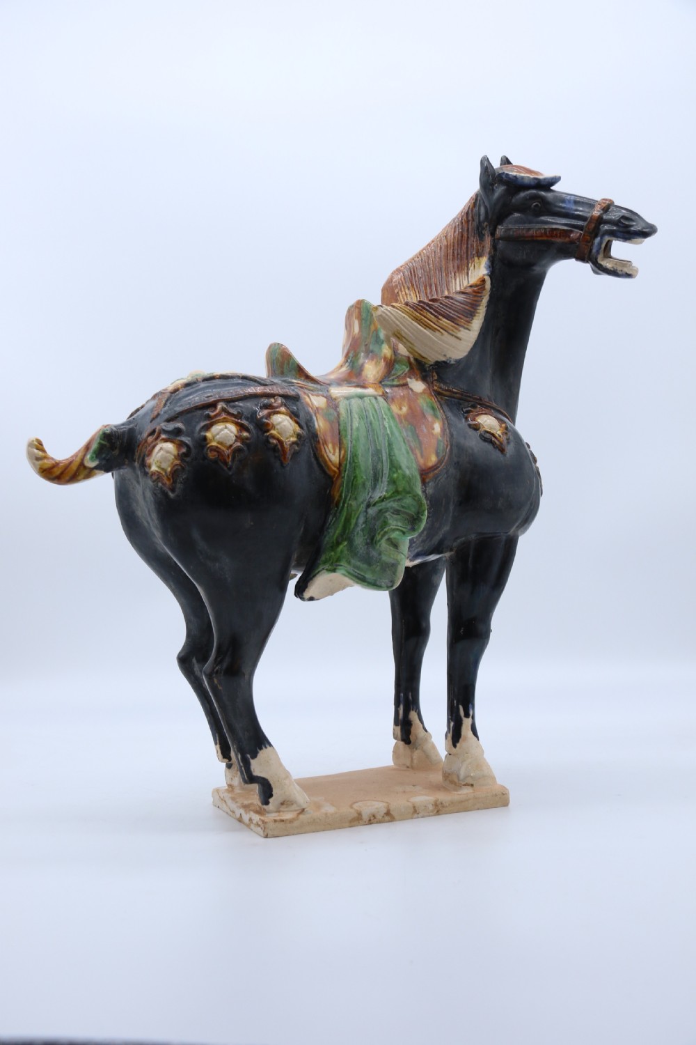 A ceramic Tang Dynasty style horse statue featuring a dark body glaze, amber mane, green draped saddle, and cream base. View XIV