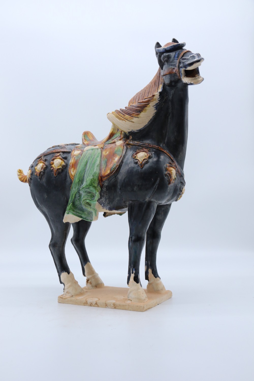 A ceramic Tang Dynasty style horse statue featuring a dark body glaze, amber mane, green draped saddle, and cream base. View XV