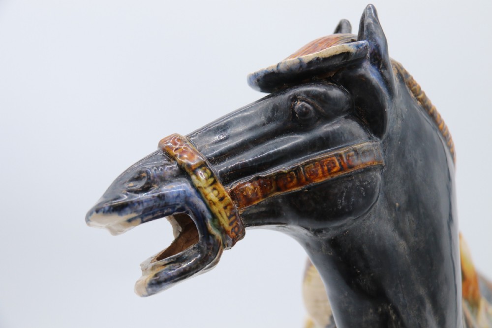 A ceramic Tang Dynasty style horse statue featuring a dark body glaze, amber mane, green draped saddle, and cream base. View XVIII
