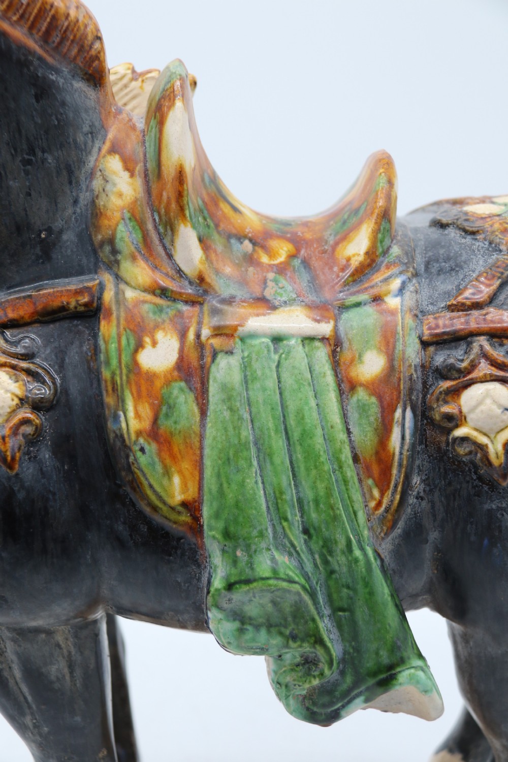 A ceramic Tang Dynasty style horse statue featuring a dark body glaze, amber mane, green draped saddle, and cream base. View XIX