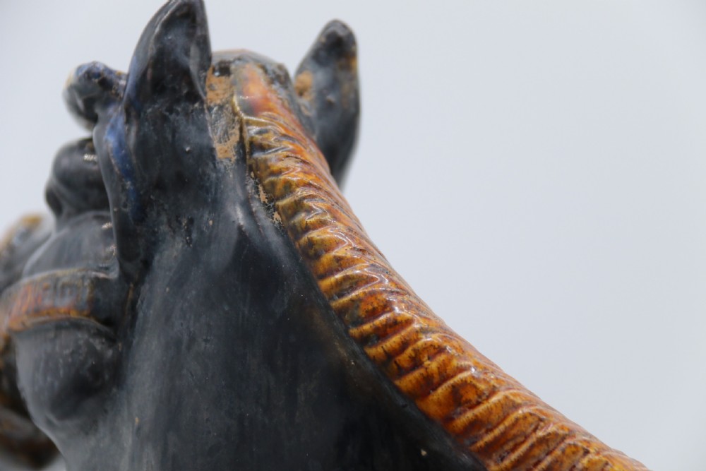 A ceramic Tang Dynasty style horse statue featuring a dark body glaze, amber mane, green draped saddle, and cream base. View XX