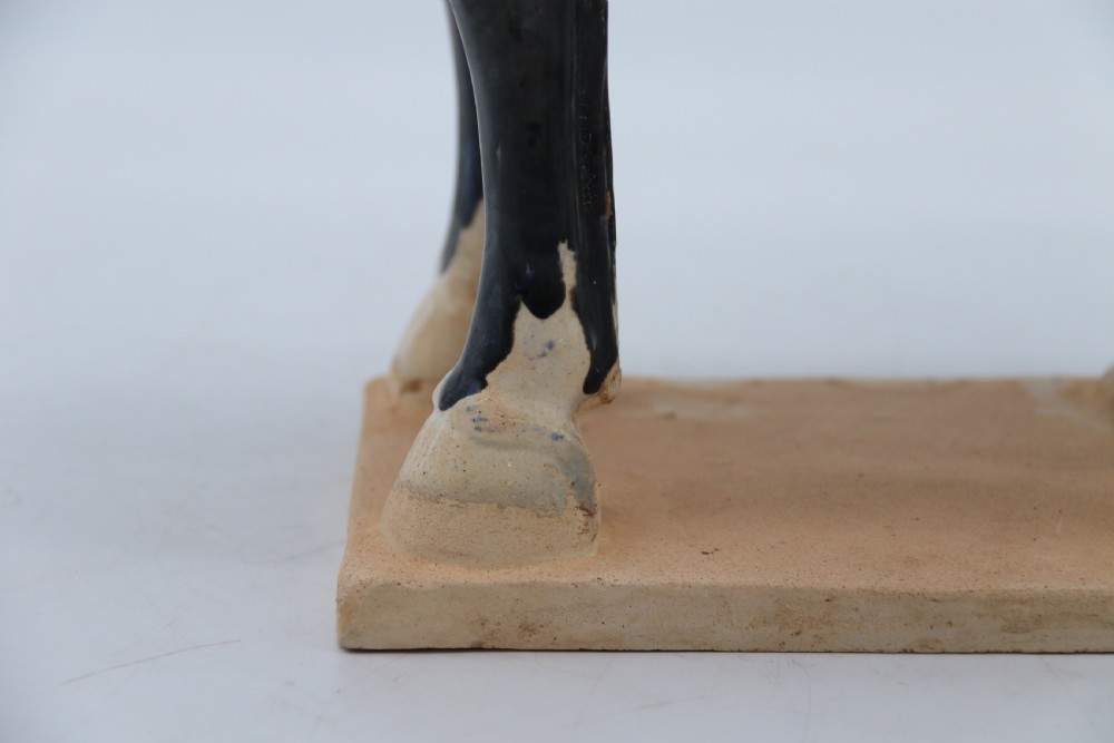 A ceramic Tang Dynasty style horse statue featuring a dark body glaze, amber mane, green draped saddle, and cream base. View XXII