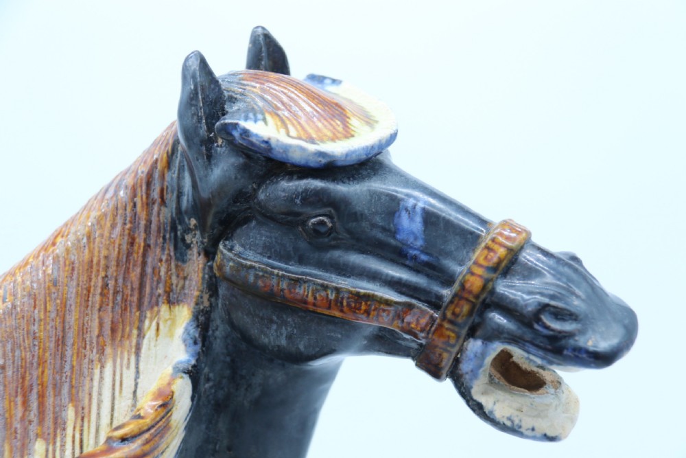 A ceramic Tang Dynasty style horse statue featuring a dark body glaze, amber mane, green draped saddle, and cream base. View XXIII