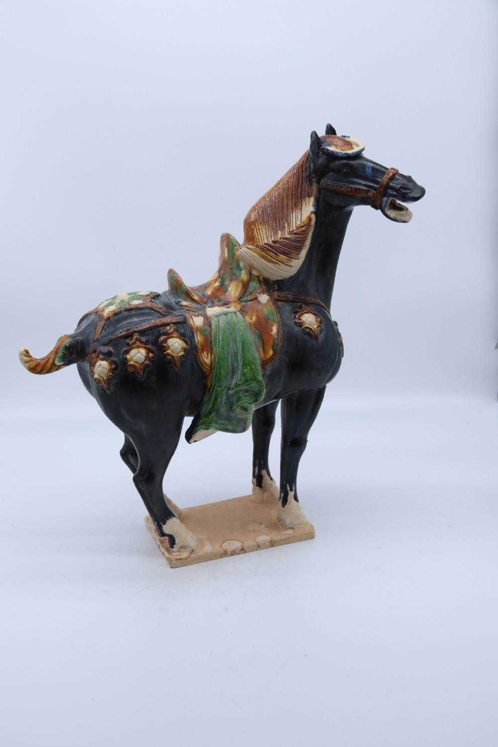A ceramic Tang Dynasty style horse statue featuring a dark body glaze, amber mane, green draped saddle, and cream base. View XXVI