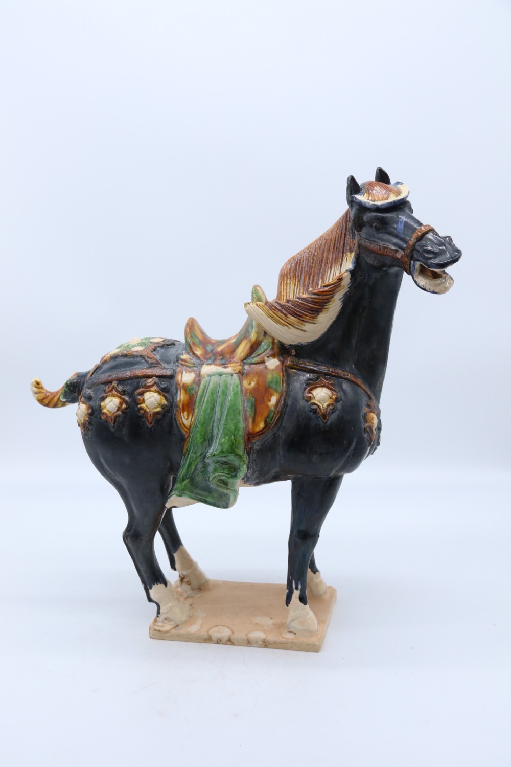 A ceramic Tang Dynasty style horse statue featuring a dark body glaze, amber mane, green draped saddle, and cream base. View XXVII