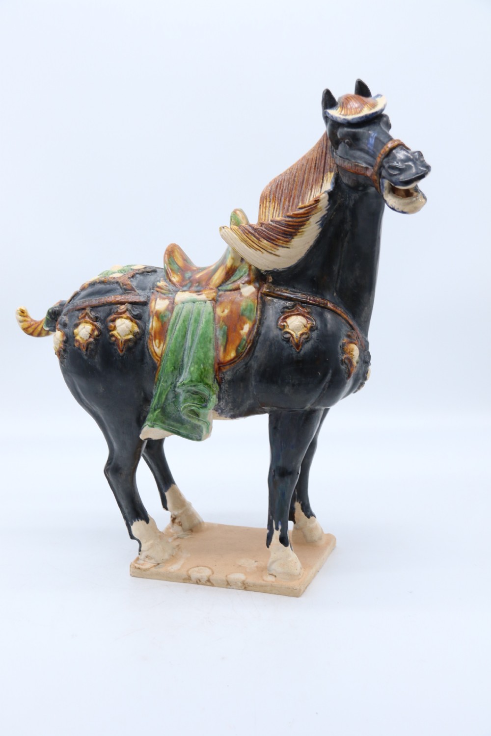 A ceramic Tang Dynasty style horse statue featuring a dark body glaze, amber mane, green draped saddle, and cream base. View XXVIII