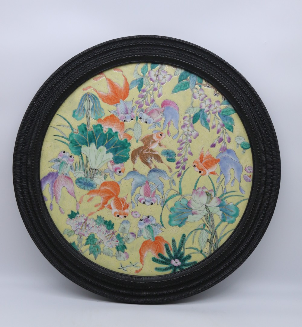 A circular framed Chinese porcelain plaque featuring vibrant hand-painted goldfish and lotus flowers on a yellow background enclosed in a carved dark frame.