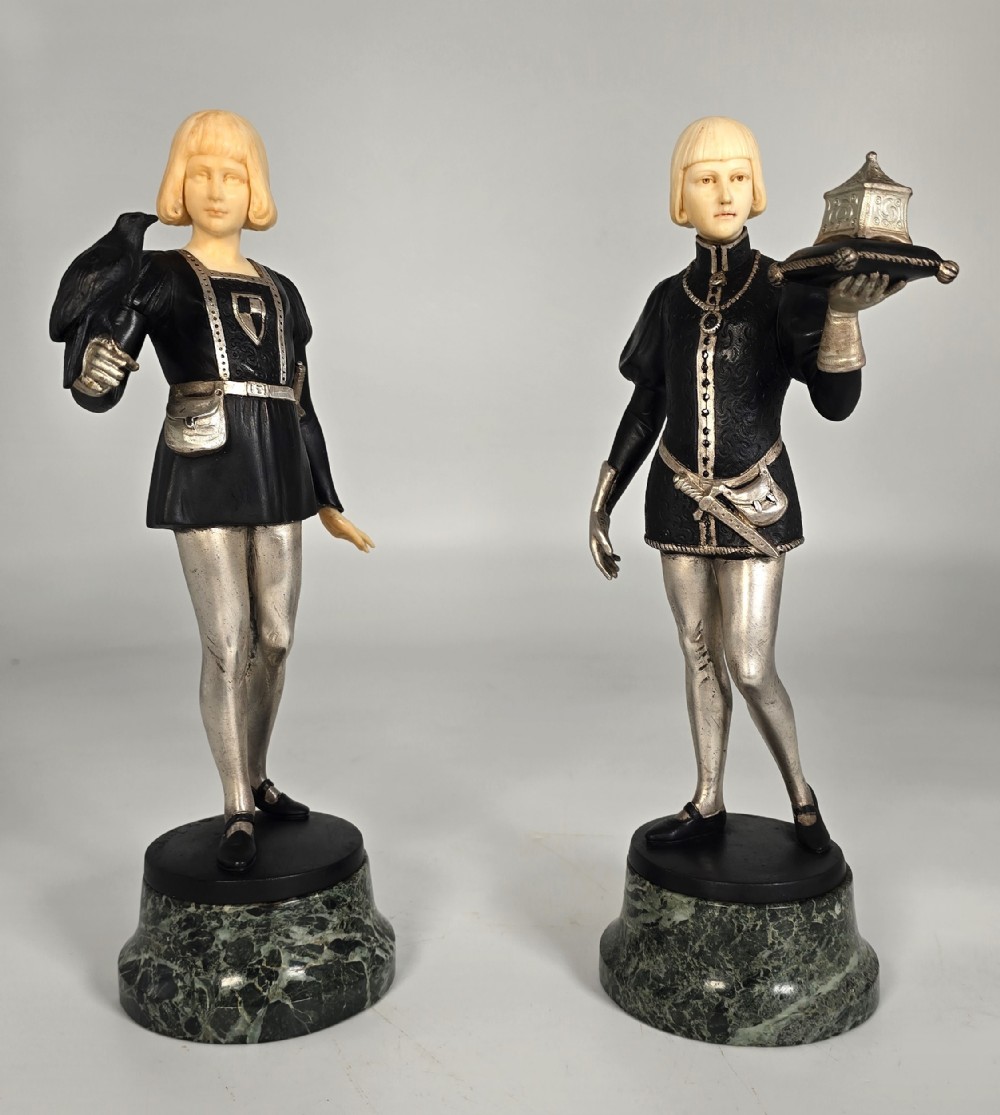 A pair of Art Deco chryselephantine statuettes depicting medieval page boys in silvered and dark bronze with carved ivory heads on green marble bases.