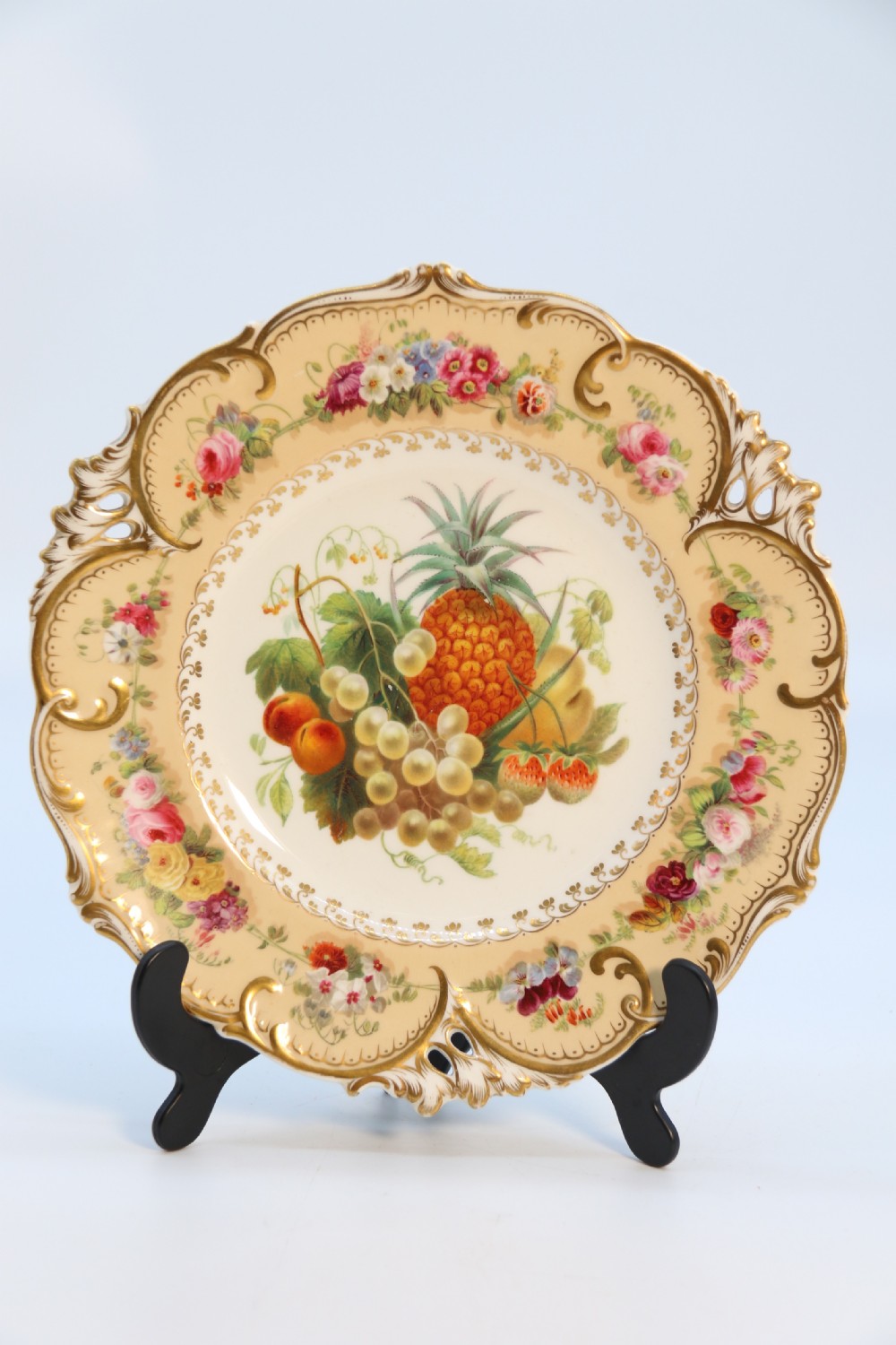 A cream-coloured circular porcelain cabinet plate featuring a scalloped gilded rim, floral swags, and a central hand-painted motif of a pineapple, grapes, and fruit.
