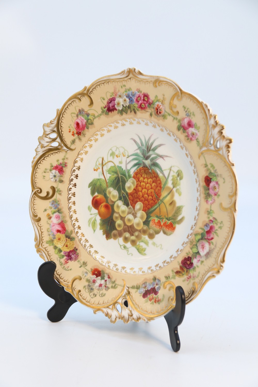 A cream-coloured circular porcelain cabinet plate featuring a scalloped gilded rim, floral swags, and a central hand-painted motif of a pineapple, grapes, and fruit. View II