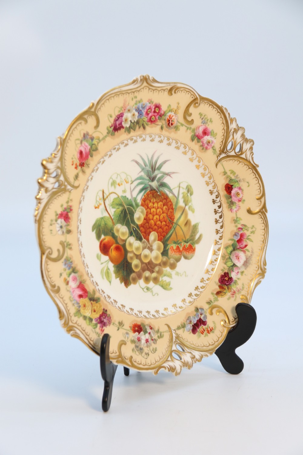 A cream-coloured circular porcelain cabinet plate featuring a scalloped gilded rim, floral swags, and a central hand-painted motif of a pineapple, grapes, and fruit. View III