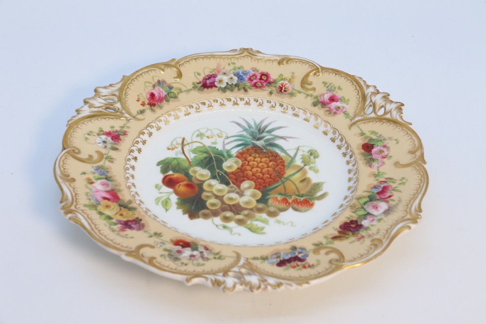 A cream-coloured circular porcelain cabinet plate featuring a scalloped gilded rim, floral swags, and a central hand-painted motif of a pineapple, grapes, and fruit. View V