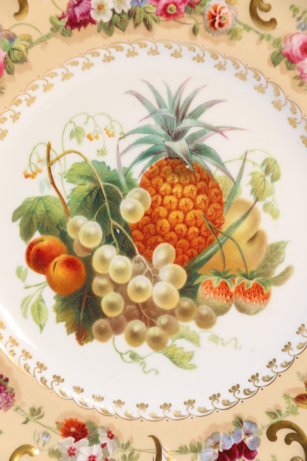 A cream-coloured circular porcelain cabinet plate featuring a scalloped gilded rim, floral swags, and a central hand-painted motif of a pineapple, grapes, and fruit. View VI