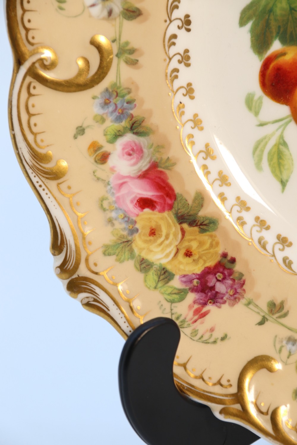 A cream-coloured circular porcelain cabinet plate featuring a scalloped gilded rim, floral swags, and a central hand-painted motif of a pineapple, grapes, and fruit. View IX