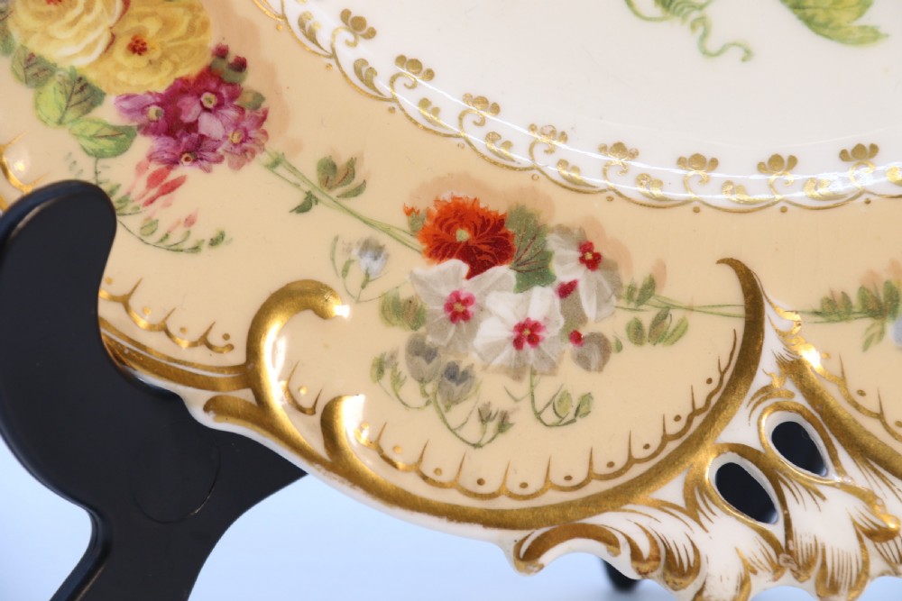 A cream-coloured circular porcelain cabinet plate featuring a scalloped gilded rim, floral swags, and a central hand-painted motif of a pineapple, grapes, and fruit. View X