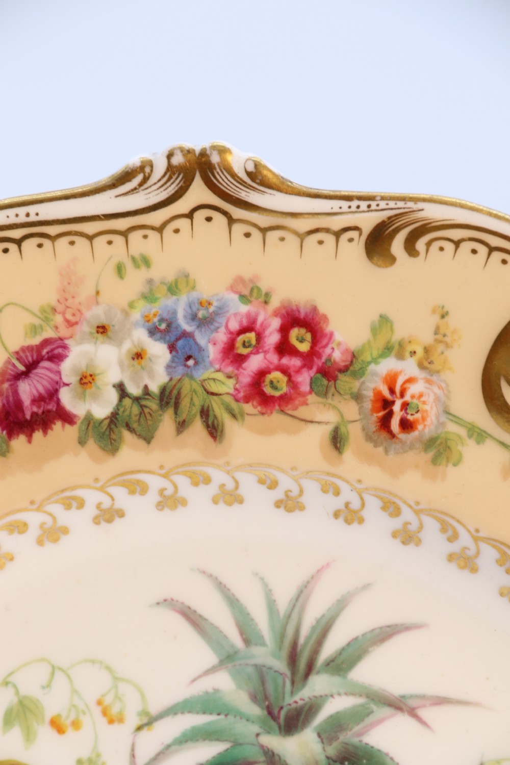 A cream-coloured circular porcelain cabinet plate featuring a scalloped gilded rim, floral swags, and a central hand-painted motif of a pineapple, grapes, and fruit. View XVI