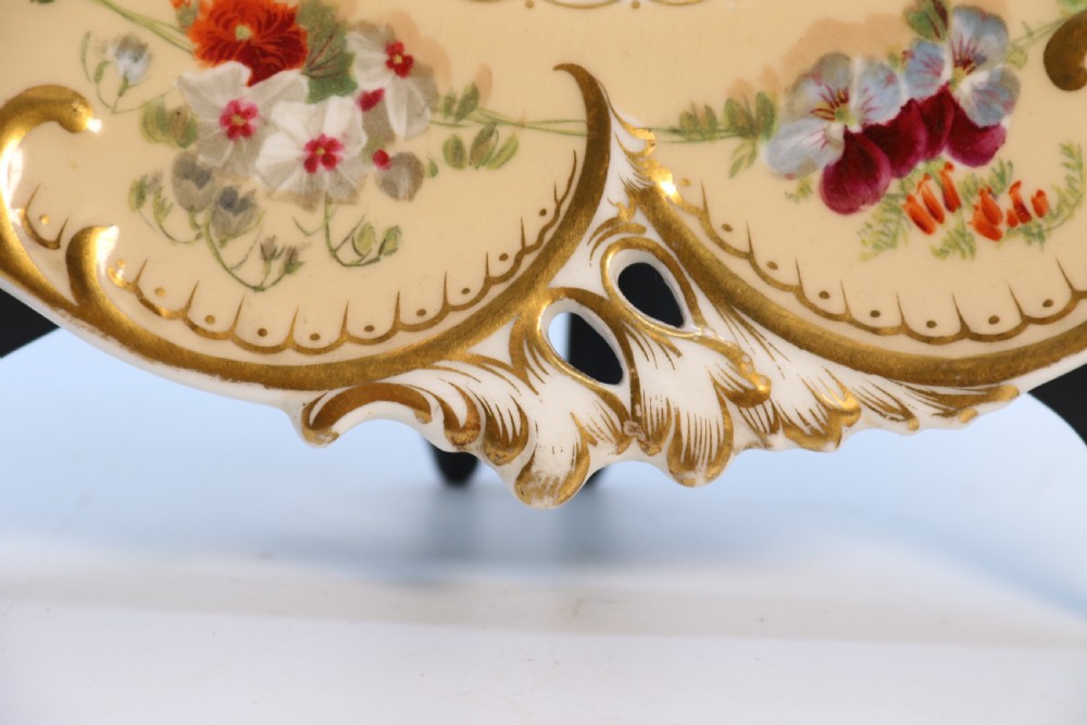 A cream-coloured circular porcelain cabinet plate featuring a scalloped gilded rim, floral swags, and a central hand-painted motif of a pineapple, grapes, and fruit. View XVII